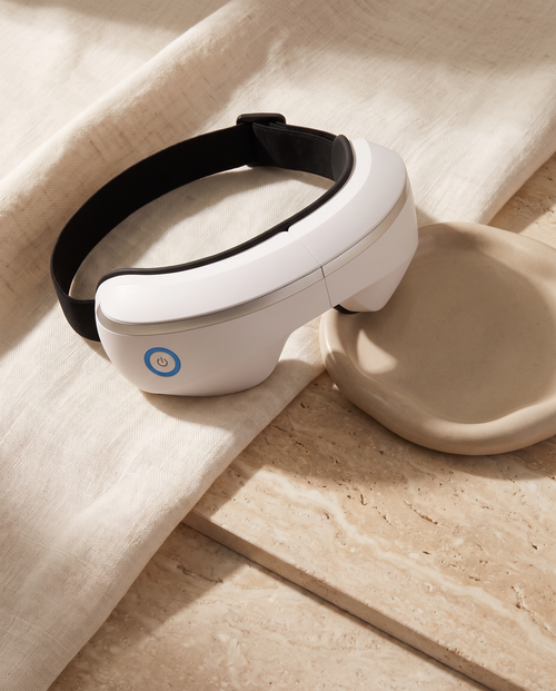 Smart Eye Massager still-life on cream linen and travertine, black head strap and power button visible