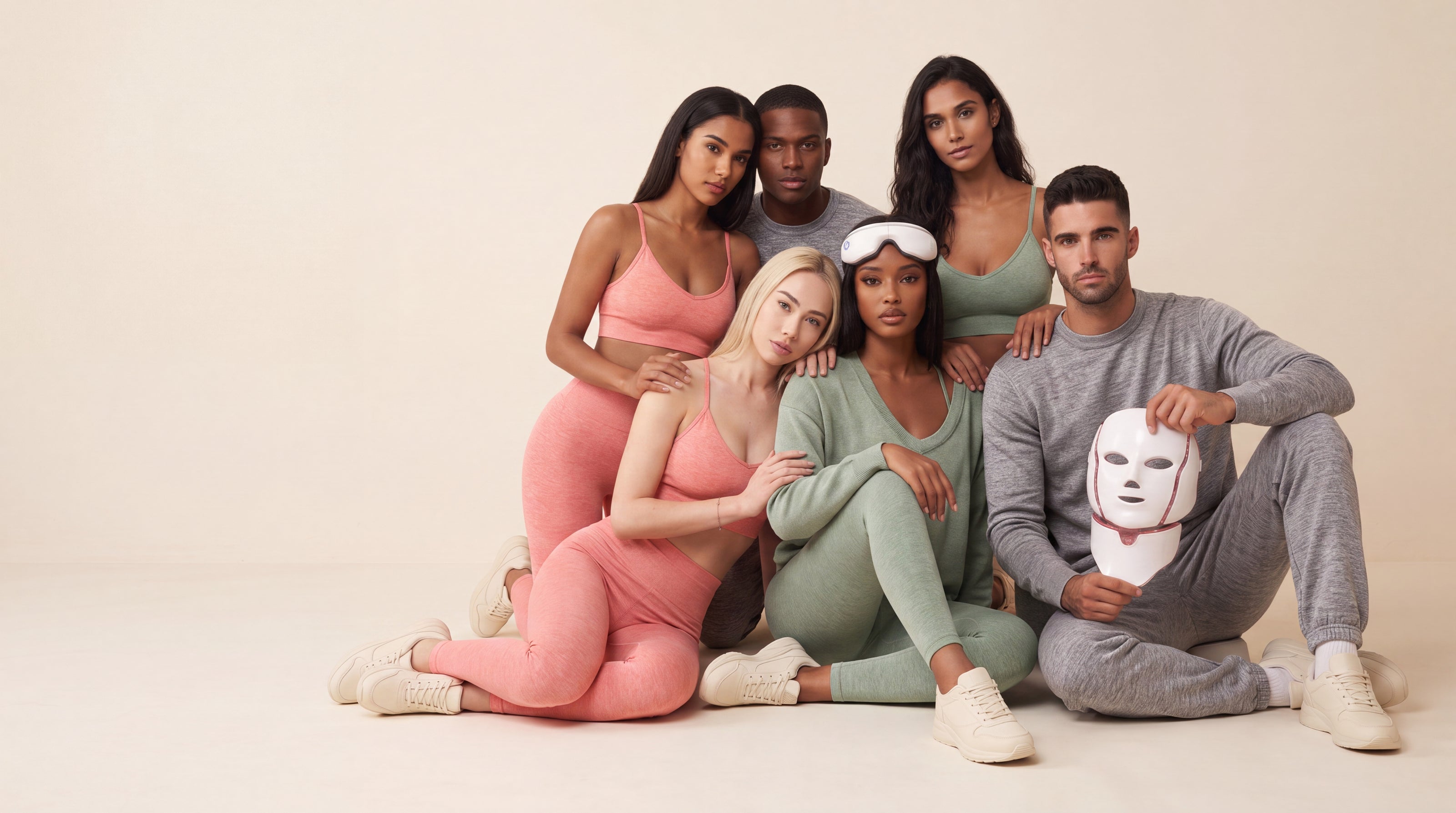 A group of six people in coral, sage and grey marl activewear on a cream studio floor, two holding the Wellness on Screen LED face mask and eye massager